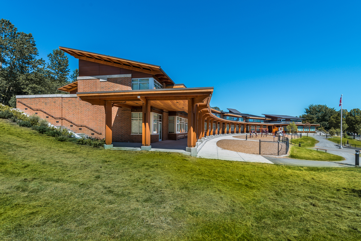 Exterior sunny daytime view of J.W. Sexmith Elementary School showing sweeping curved mass timber covered walkway along building front which features glue-laminated timber beams, parallel strand lumber and solid-sawn heavy timber