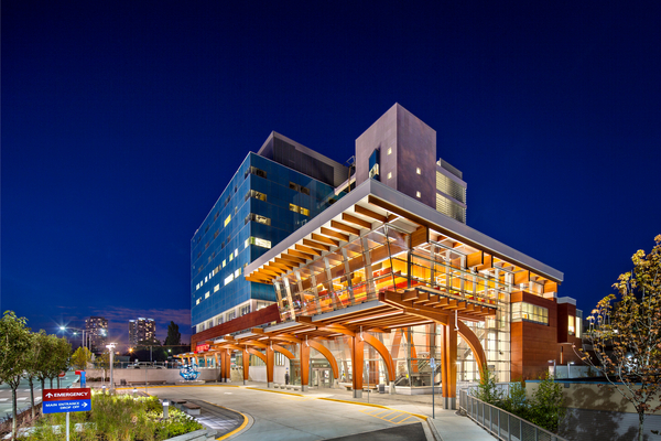 Exterior distant evening view of multi-storey glass, wood, metal Surrey Memorial Hospital Critical Care Emergency Services Entrance showing curved glue-laminated timber (Glulam) columns, glulam beams, and decorative millwork